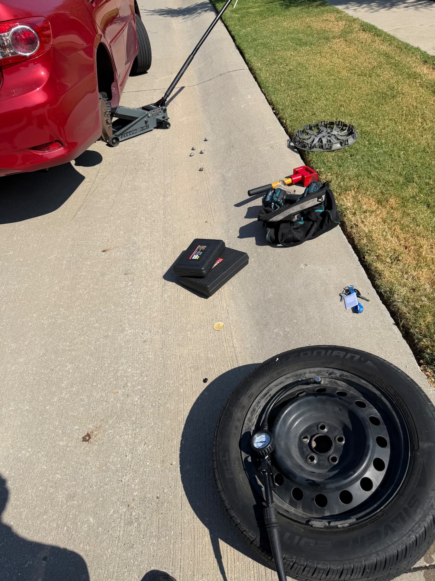 Spare tire change in Denton, TX 76201 Red sedan lifted for a roadside tire change with the removed wheel, compact spare, pressure gauge and tools in Denton, Texas 76201