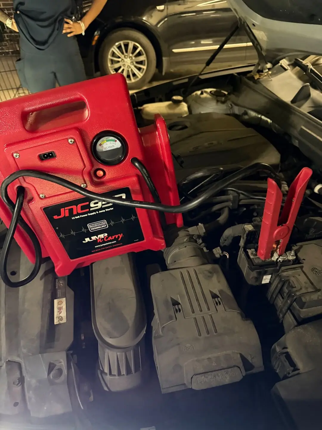 Red jump pack clamp attached to the positive terminal of a car battery in an open engine bay during an Arlington, Texas roadside jump start at night