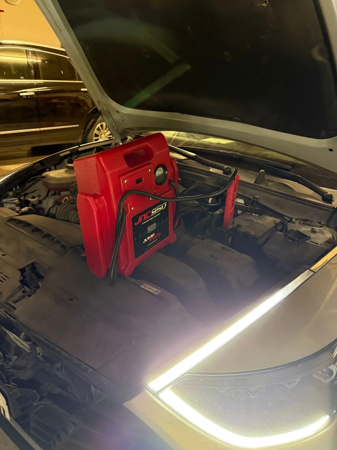Red JNC 950 professional 12-volt jump pack sitting in the open engine bay of a silver sedan during an Arlington, Texas roadside jump start