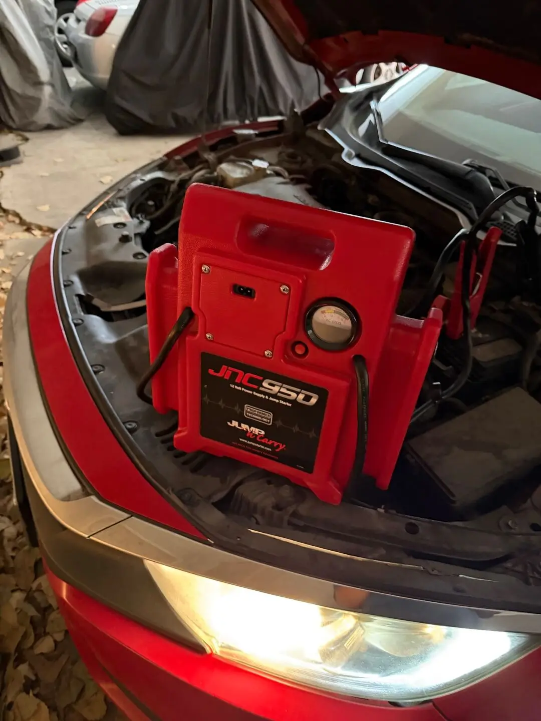 Red JNC 950 professional 12-volt jump pack clamped to a red car battery with the hood open during a Dallas, Texas 75231 roadside jump start, headlight lit
