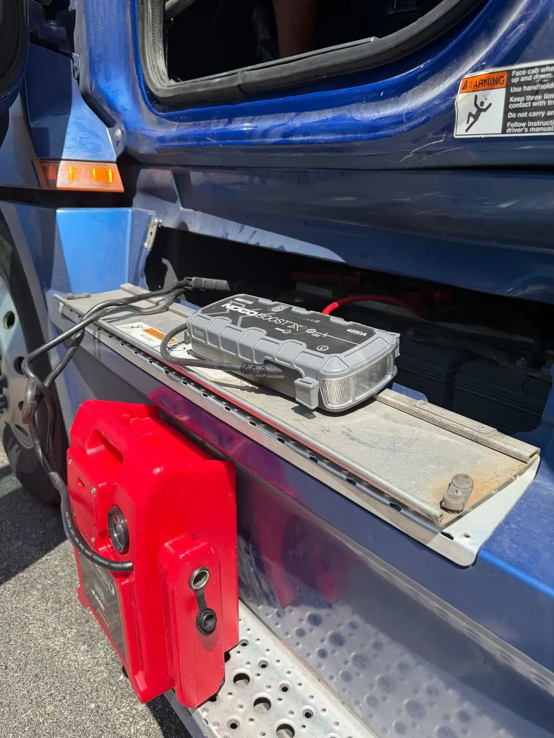 Semi truck jump start in downtown Dallas, TX 75204 — 4250 amp commercial jump pack NOCO Boost X 4250 amp commercial jump pack sitting on the frame step of a blue semi truck next to the battery box during a downtown Dallas, Texas 75204 jump start