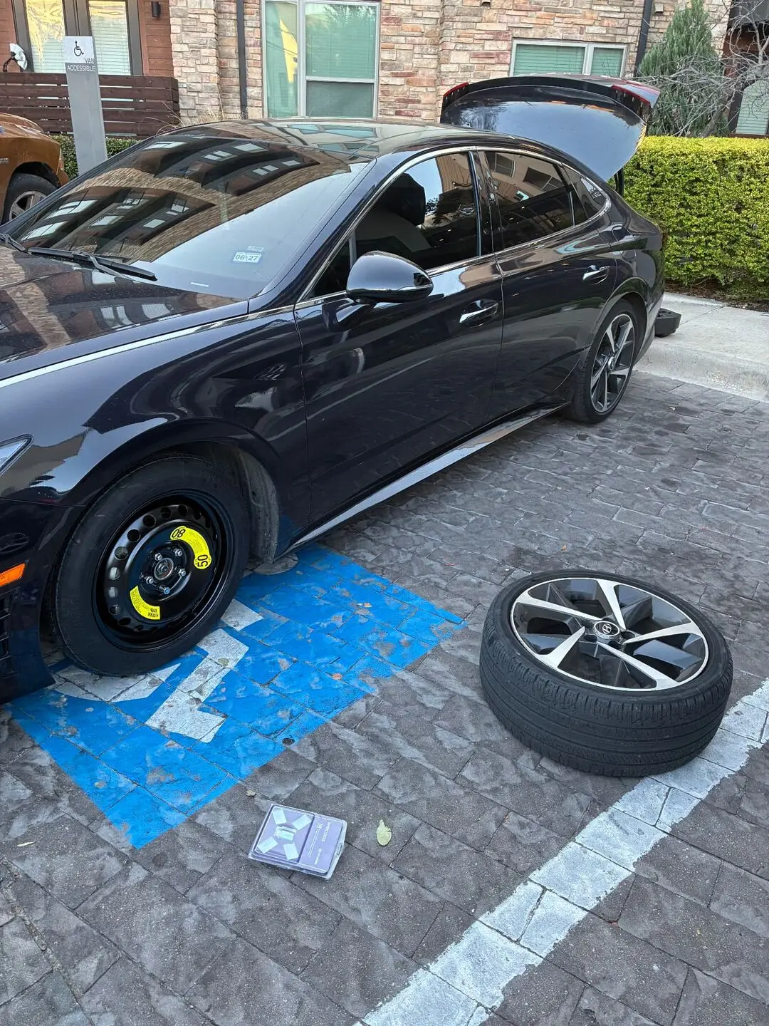 Black Hyundai Sonata with a donut spare tire mounted in an Allen, Texas 75013 apartment parking space and the removed alloy wheel on the pavers