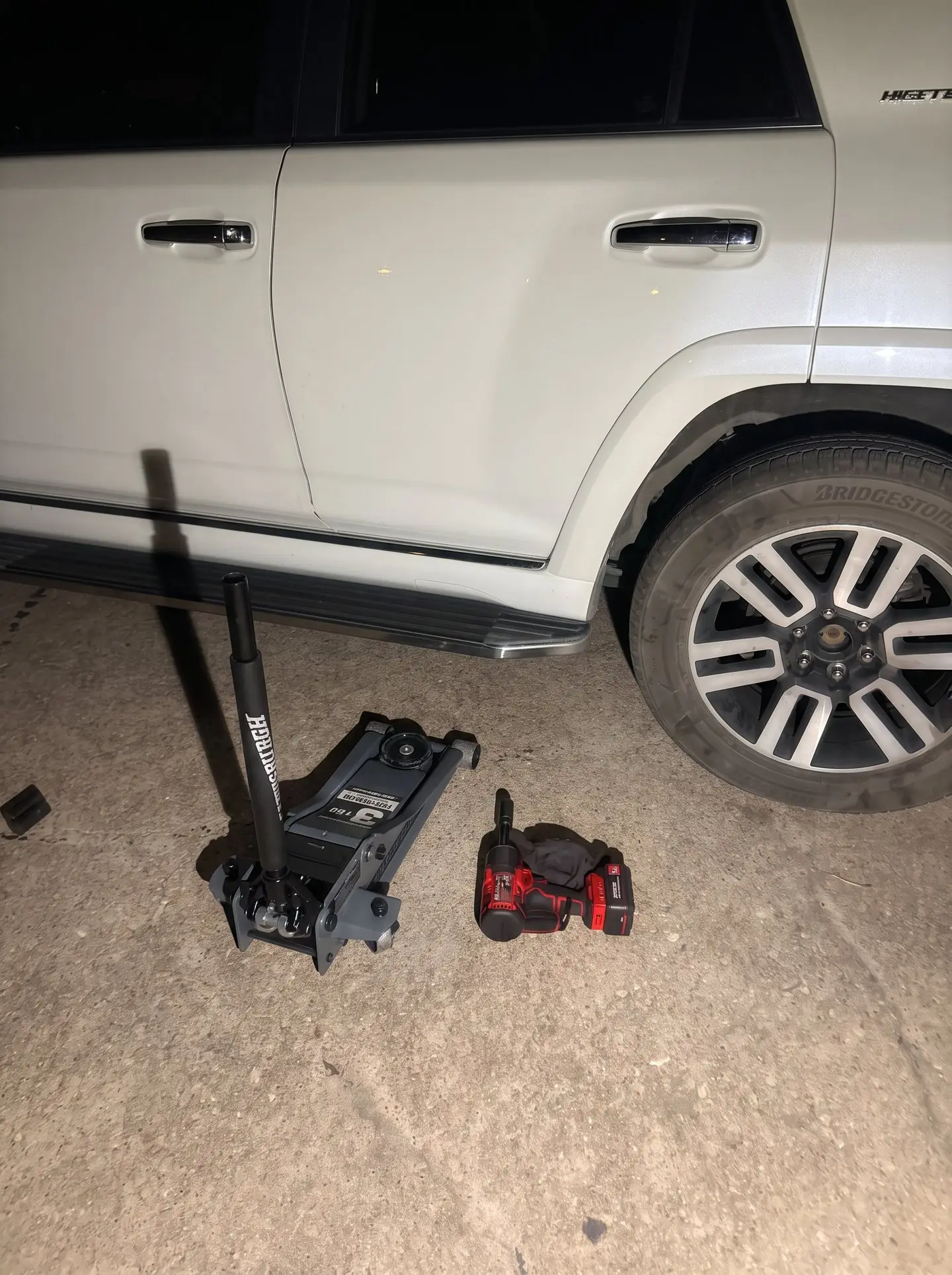 White SUV raised on a low-profile floor jack at night during an after-hours flat tire change in Carrollton, Texas, with a cordless impact wrench on the pavement