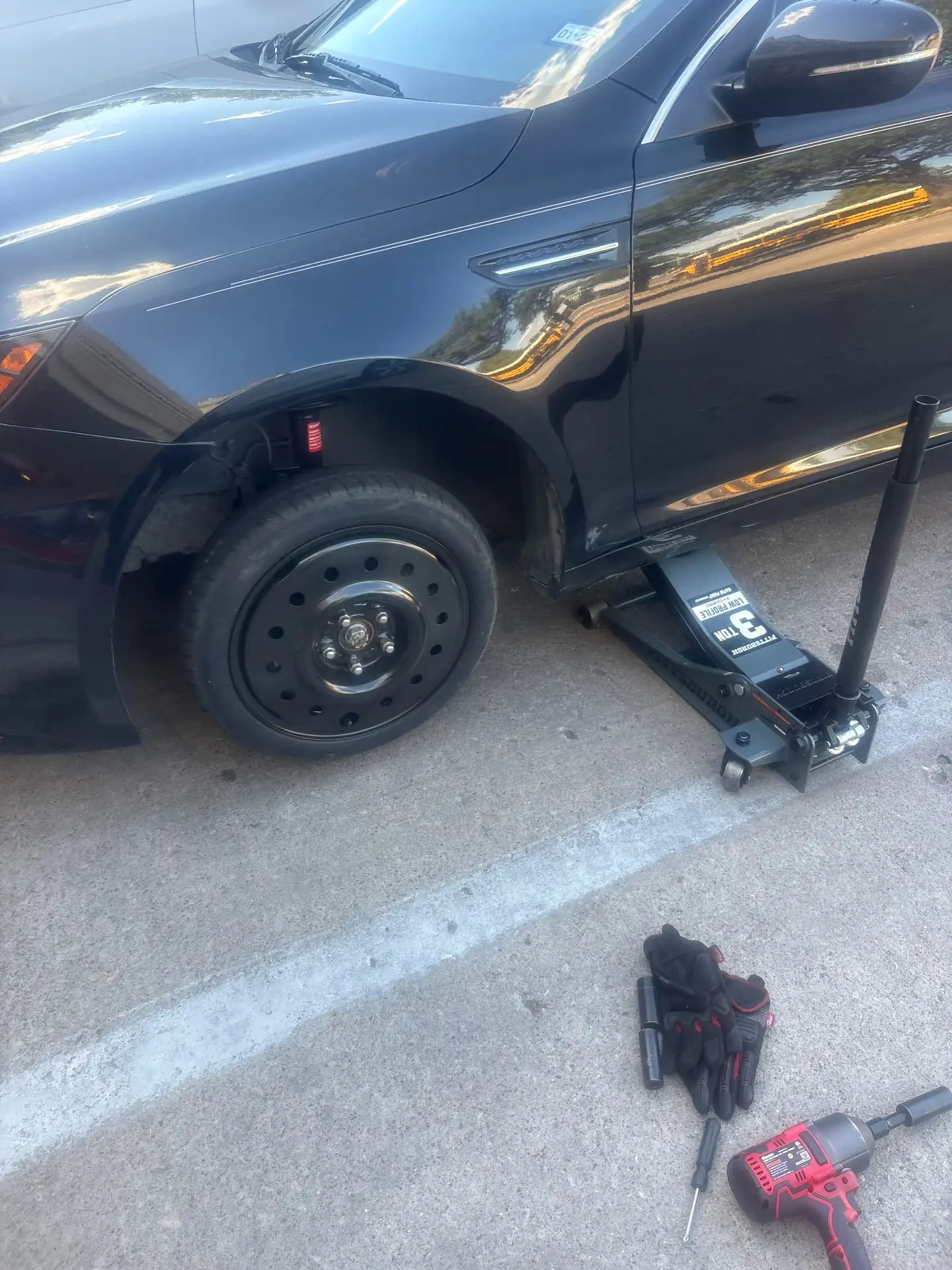Highway 35 Roadside technician changing a flat tire on a black sedan lifted with a floor jack on a Dallas street