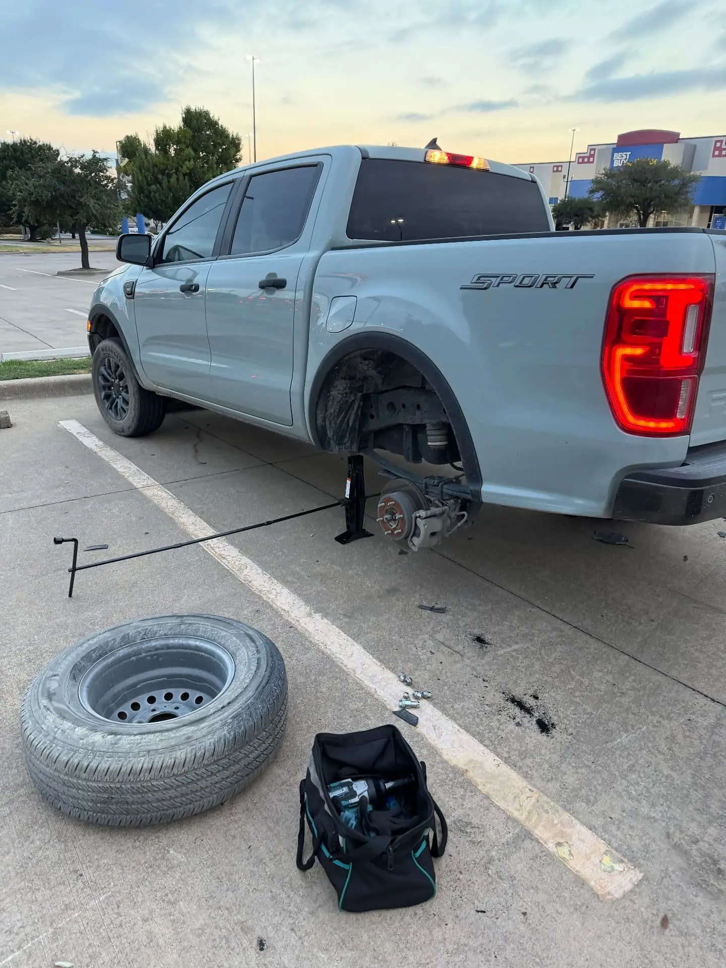 Tire Change in Dallas, TX — Highway 35 Roadside Ford Ranger Sport pickup raised on a jack with the rear wheel removed during a Dallas, Texas roadside flat tire change, spare tire and tool bag on the pavement