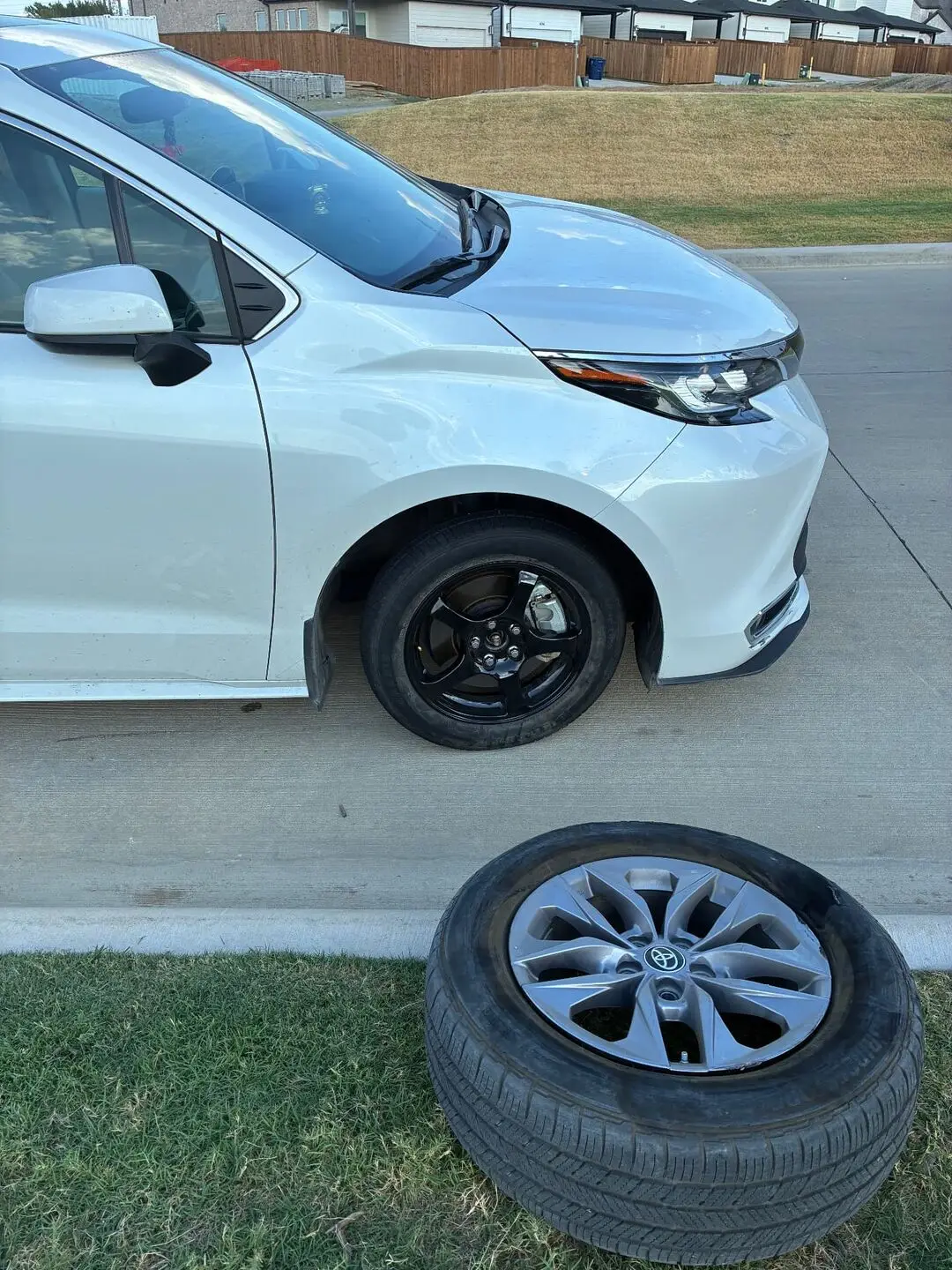 Spare tire change on a Toyota Sienna in Frisco, TX 75034 White Toyota Sienna at a Frisco, Texas 75034 curb with a black steel spare wheel mounted and the removed flat tire resting on the grass