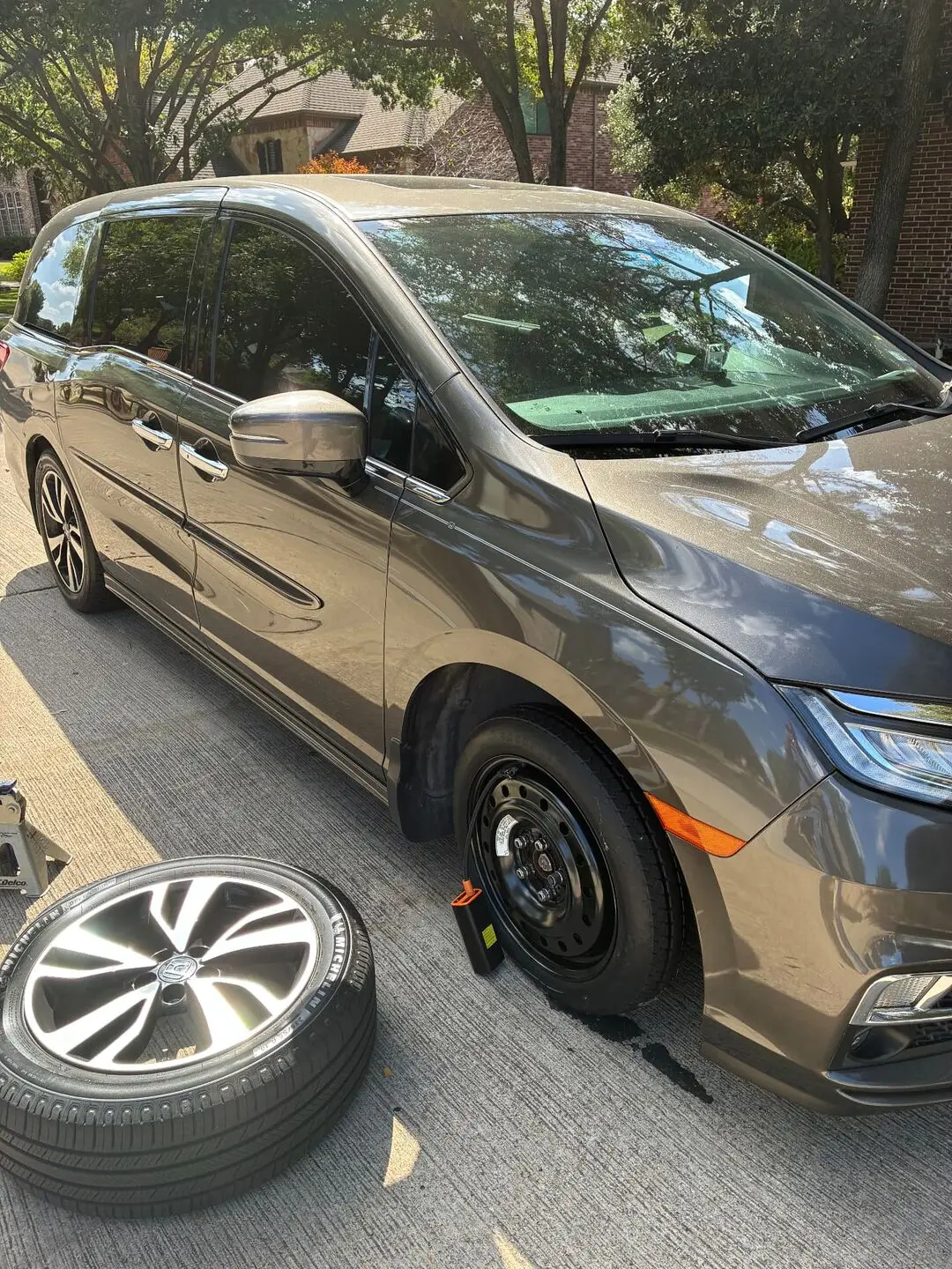 Spare tire change on a Honda Odyssey in McKinney, TX 75070 Compact spare mounted on a grey Honda Odyssey in a McKinney, Texas 75070 driveway with the removed flat alloy wheel lying on the concrete