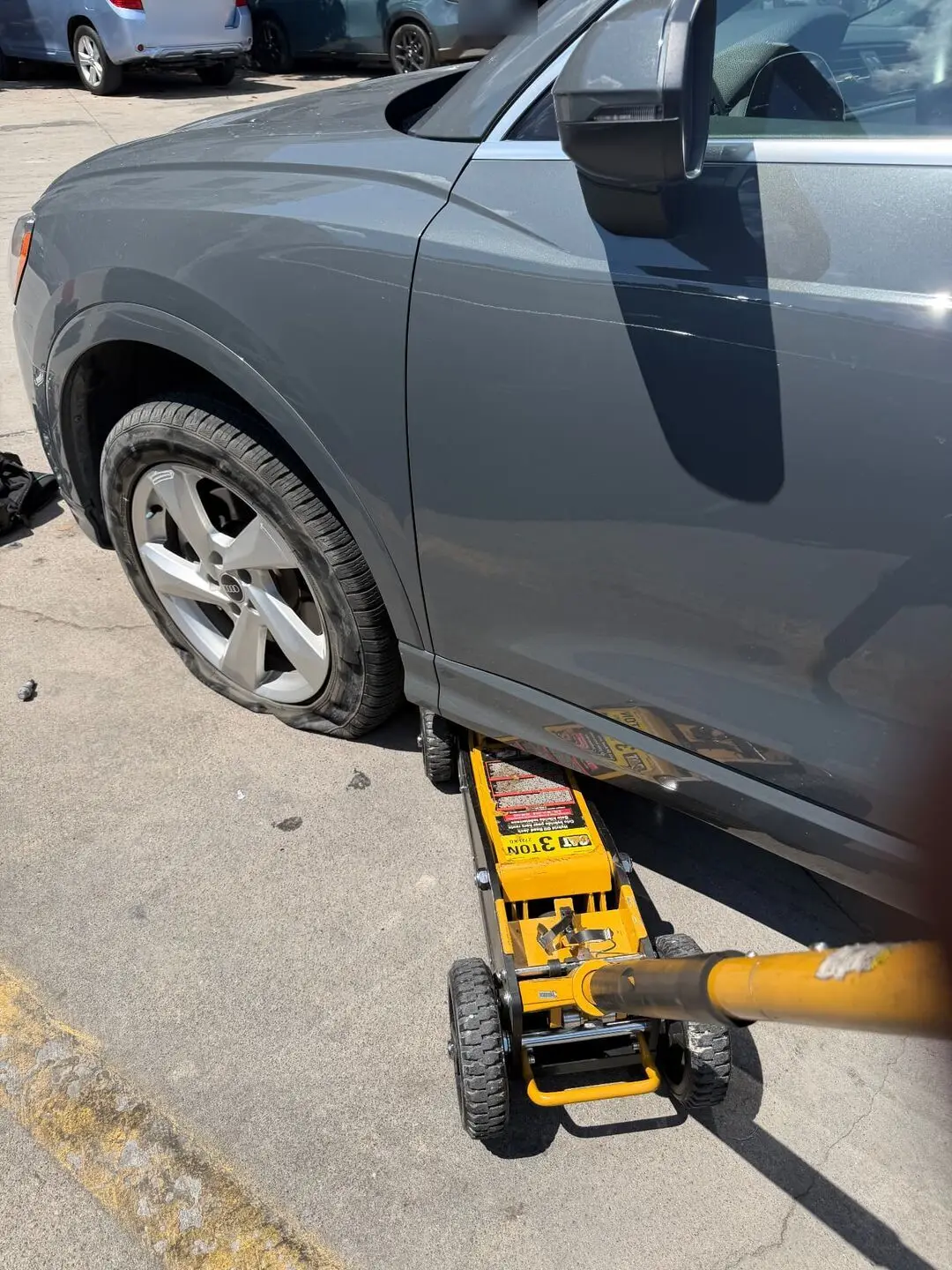 Grey Audi Q3 raised on a yellow 3-ton floor jack with a flat front tire during a mobile tire change on the SMU campus in Dallas, Texas 75205
