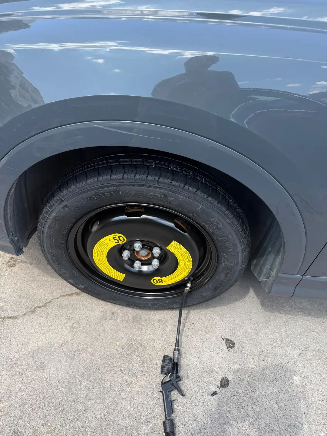 Compact temporary spare tire mounted on a grey Audi Q3 and being inflated with an air hose after a mobile tire change on the SMU campus in Dallas, Texas 75205